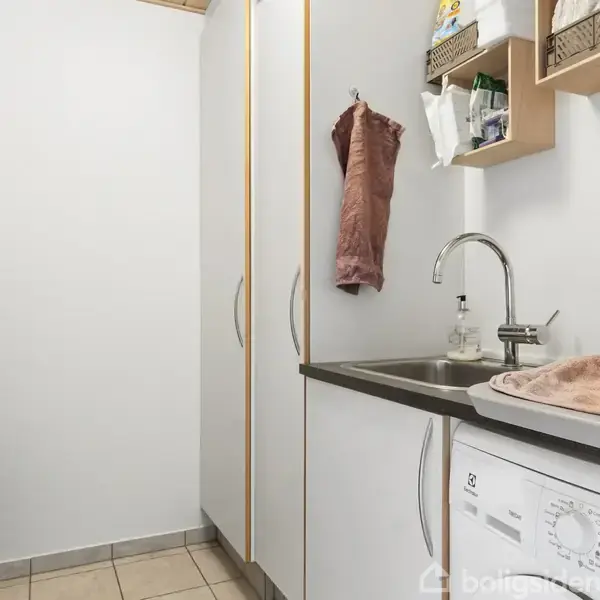 Et vaskerum med en hvid vaskemaskine under en foldet lyserød håndklæde placeret ved siden af en vask. Håndklæde hænger på væggen, med skabe og hylder omkring.