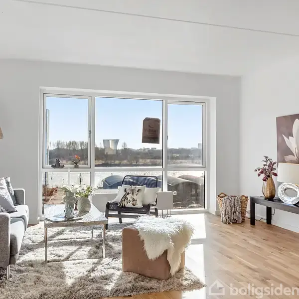 Sofa med puder står i en stue med store vinduer, der lukker sollys ind. Stuen er indrettet med gulvlampe, sofabord, tæppe og dekorativt bord.