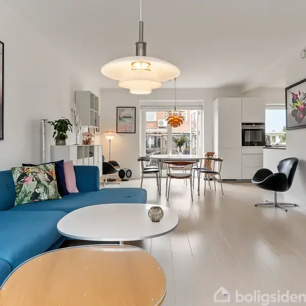 Blå sofa i moderne stue med farverige kunstværker op ad væggen. Rundt sofabord foran sofaen. Spisebord med stole ses i baggrunden ved et køkken. Lyst trægulv.