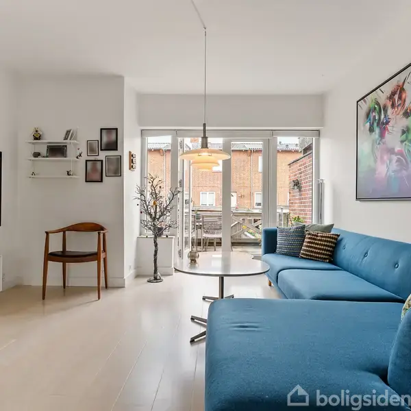 En blå sofa står i en moderne stue med trægulv, fladskærms-TV på væggen, kunstmaleri og spisebord ved vinduet. Indretningen er lys og skandinavisk med udsigt til en altan.