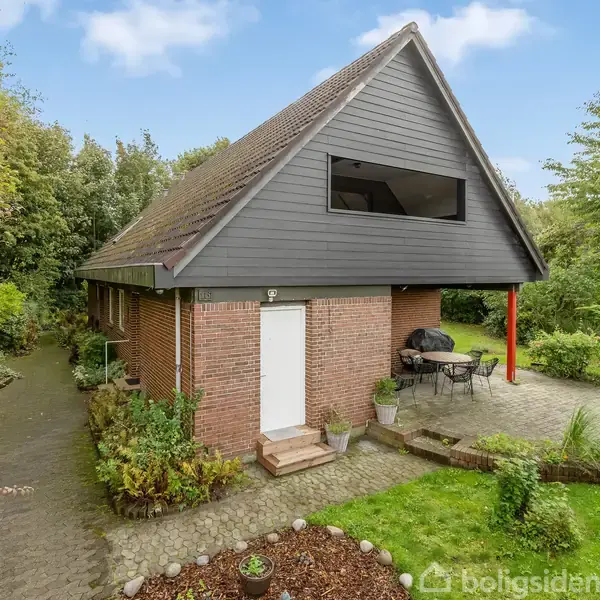 Et to-etagers hus med muret vægge og stejlt tag står omgivet af frodig have med stier, buske og træer. Der er en terrasse med bord og stole foran huset.