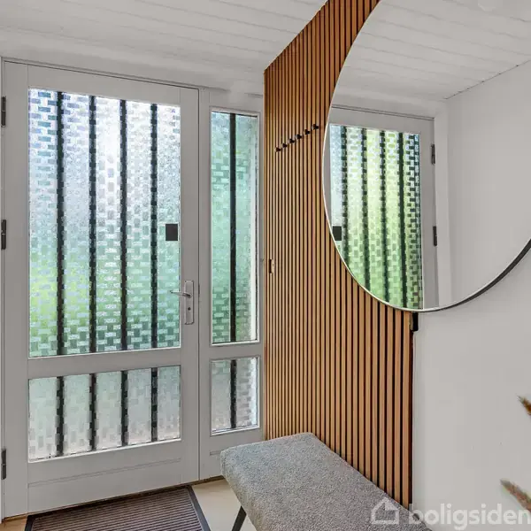 Stor, rund spejl hænger på væggen ved siden af en bænk i en lys gang med glasdør og matterede vinduer. Tynde trælameller skaber en elegant ruminddeling.