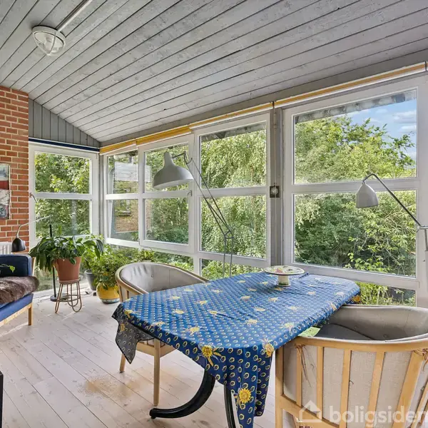 Veranda med bord dækket med blåblomstret dug, omgivet af stole. Planter i potter og naturligt lys fra store vinduer med udsigt til frodige træer. Maleri hænger på murstensvæggen.