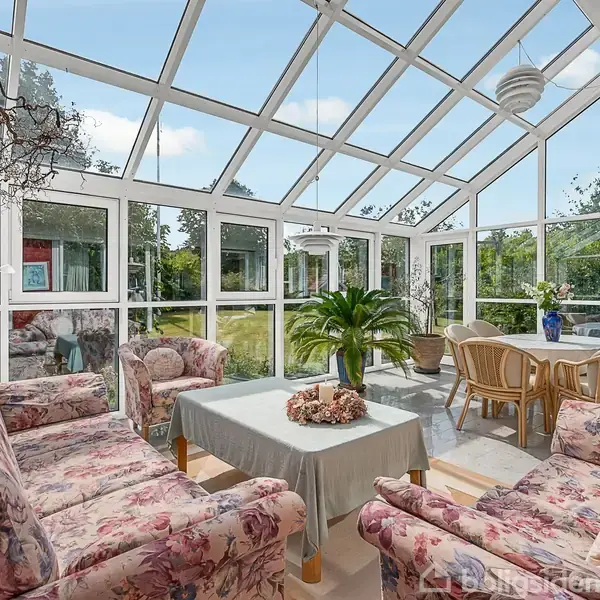 Blomstrede sofaer står i en glaskonstruktion, hvor de omkranser et bord med en plante. Ved siden af er et spisebord med stole. Udenfor ses grønne træer og en blå himmel.