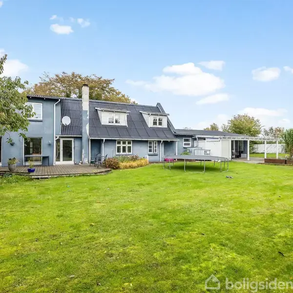 Et stort hus med flere vinduer står stille i en velplejet have med grøn plæne, omkranset af træer og buske under en blå himmel. Tekst: ingen.