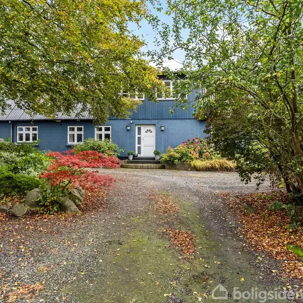 Blåt hus står stille omkranset af farverige buske og træer. Grusvej leder op til hoveddøren i et frodigt, grøn haveområde.