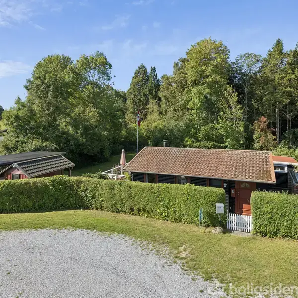 Et lille hus med rødt tag står omgivet af tæt hæk. I baggrunden er der frodig skov og nogle mindre strukturer. Himlen er klar med få skyer.