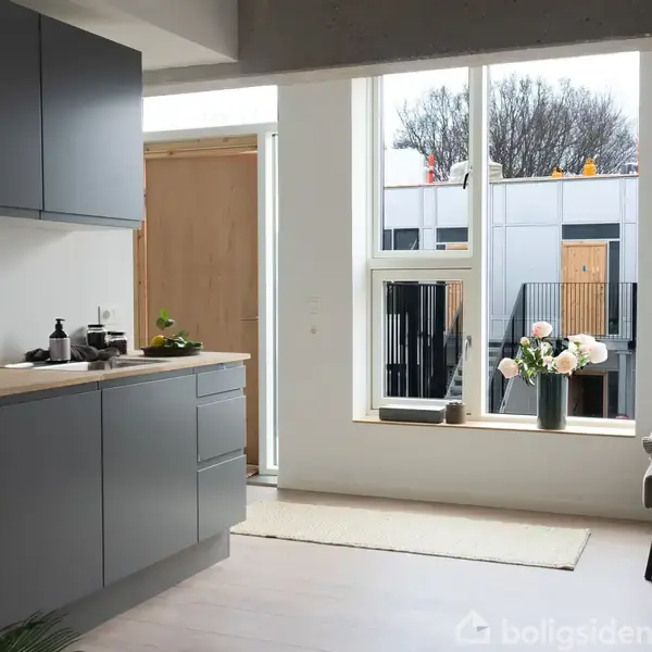 Et moderne køkken i neutrale farver med grå skabe og indbygget ovn. En stor vase med blomster står i vindueskarmen. Udenfor ses moderne boligbygninger og træer.