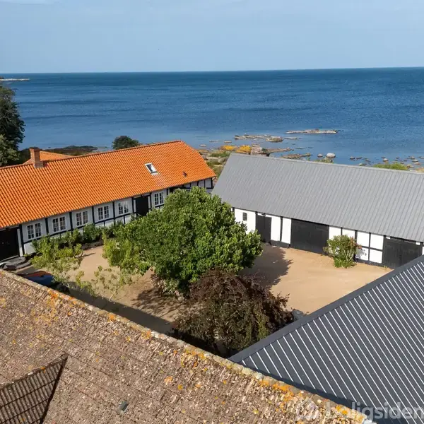 Gårdhus med rødt tegltag og sort-hvide bindingsværkshuse omringer en gårdsplads med træer, beliggende ved havet på en solrig dag.