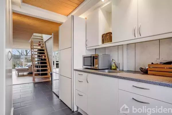 Køkken med hvidt skabsarrangement og grå bordplade, indeholder mikroovn og køkkenredskaber. Trappe fører til opholdsstue med sofa i baggrunden. Lys træloft skaber en varm atmosfære.