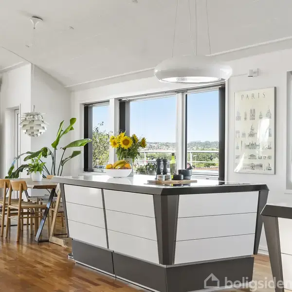 Et moderne køkken med hvidt køkkenbord, på bordet står en vase med solsikker. Ved siden af er et spisebord med stole. Store vinduer giver udsigt til en terrasse.