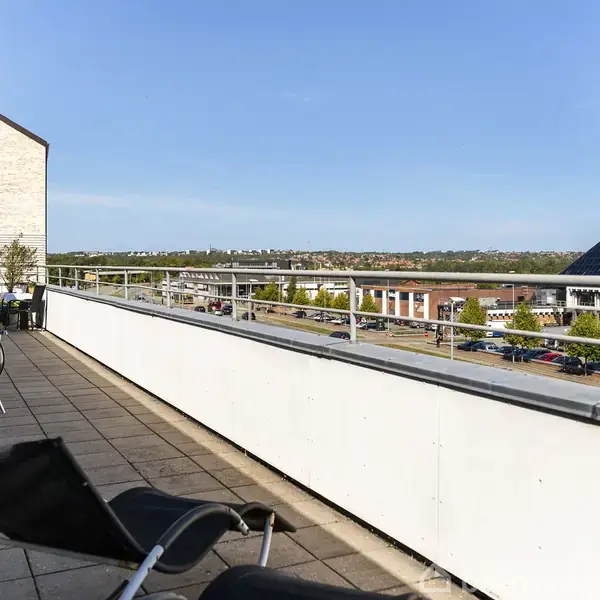 En balkon med hvide rækværk og sorte stole står tom, mens grillen er stillet til side. Udsigten viser en by med lave bygninger under en klar blå himmel.