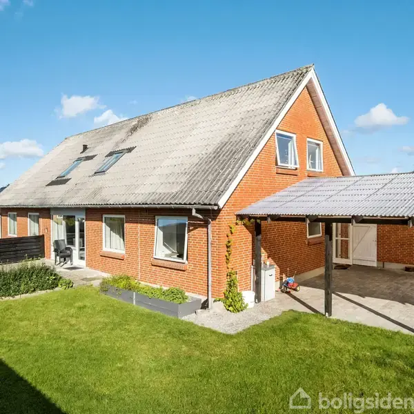 Rødt murstenshus med skråtag, carport til højre, omgivet af velholdt græsplæne og blå himmel med skyer.