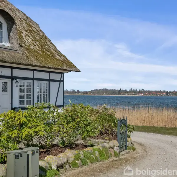 Stråtækt traditionelt hus med hvid facade og sorte bindingsværk detaljer, er placeret ved en grusvej tæt på en sø med siv og fjern udsigt til byområde. Tekst "5".