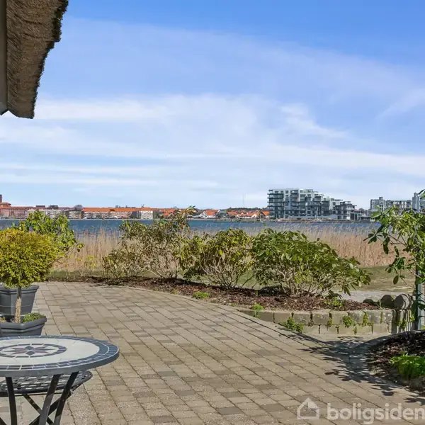 Et rundt bord med stole står på en flisebelagt terrasse ved siden af et hus. Udsigten viser en sø omgivet af bebyggelse og blå himmel i baggrunden.