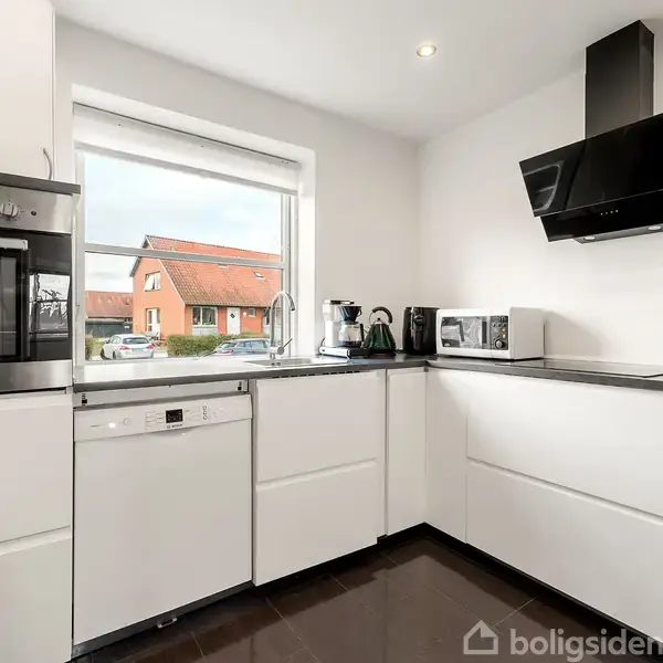 Et moderne køkken med hvide skabe og sorte bordplader. En ovn, opvaskemaskine og emhætte er synlige. Udenfor vinduet ses rødt tag og huse. Frugtskål står på bordet.