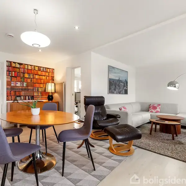 Et rundt spisebord med seks stole står midt i et lyst rum med en biblioteksvæg i baggrunden. En hvid sofa og lænestol er placeret ved siden af.