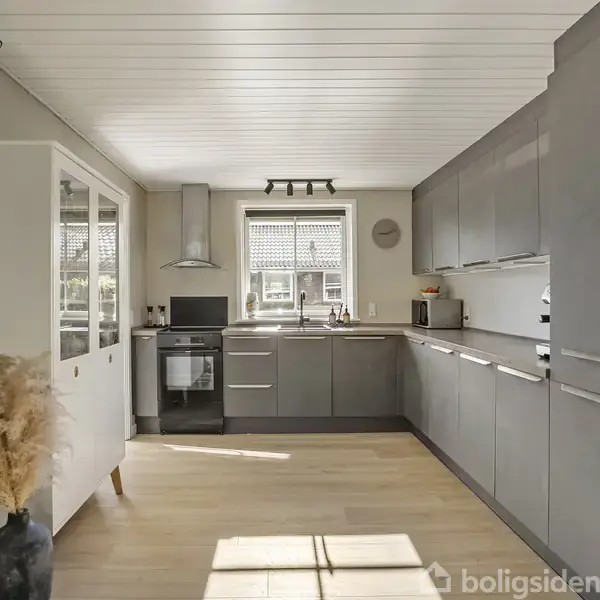 Køkken med grå skabe og en indbygget ovn. Sollys skinner ind gennem vinduet, der er placeret over køkkenbordet, og lyser op på det lyse trægulv.