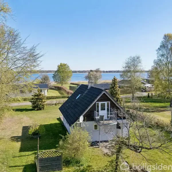 Et lille hus står stille i en frodig have med høje træer. I baggrunden ses en sø under en klar blå himmel.