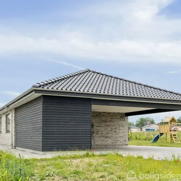 En moderne carport med fladt tag står ved siden af et enkelt-etagers hus. Omkringliggende græsplæne har en legeplads i baggrunden under en let skyet himmel.