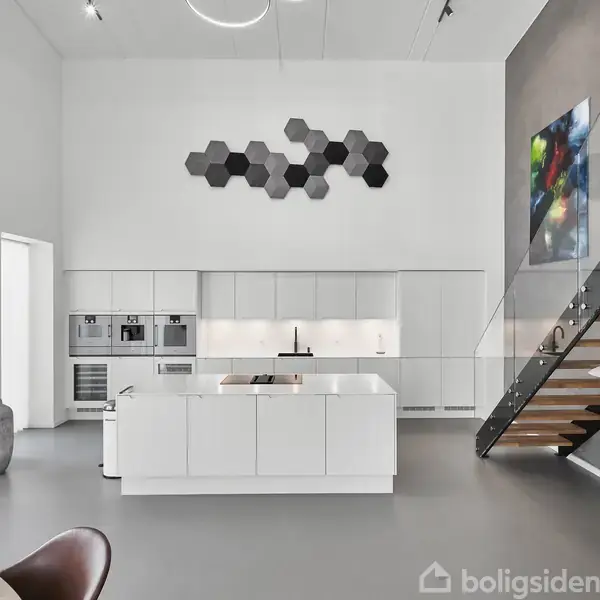 Et moderne køkken med en stor, hvid ø står centralt. På væggen hænger hexagonformet kunst. Til højre ses en glastrappe, og rummet har højt til loftet og minimalistisk indretning.