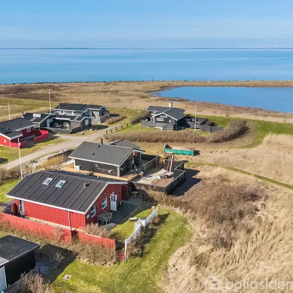 Rødt hus ligger blandt træhuse ved kysten. Husene har græsarealer omkring sig og udsigt til havet. I baggrunden ses en stor sø samt åbent vand.
