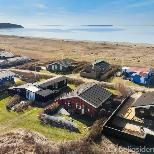 Flere sommerhuse står stille på en kystnær græsmark, med en bred sandstrand og det rolige hav i baggrunden under en blå himmel.