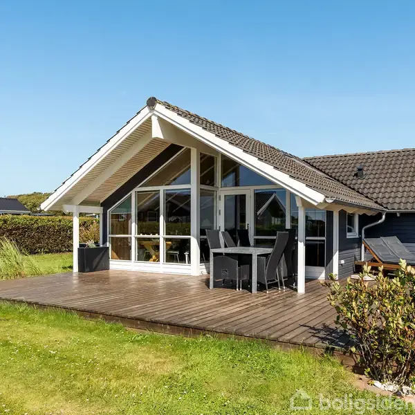 Et moderne sommerhus med store glasfacader står på en træterrasse. Havemøbler er placeret udenfor. Huset ligger i et grønt område med velholdt græsplæne og buske.