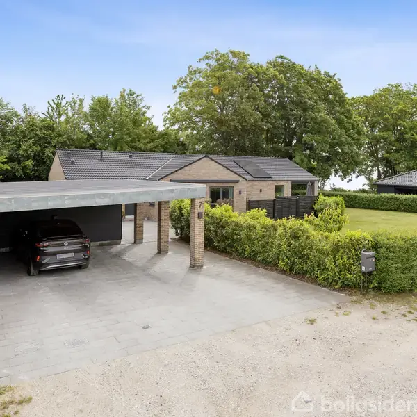 Et moderne hus med fladt tag står stille omgivet af grønne buske og træer. En carport til venstre huser en sort bil, og belægningen er fliser.