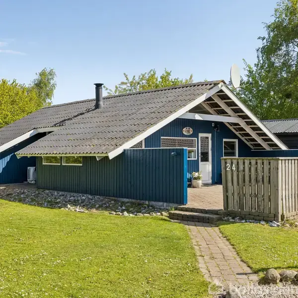 Et blåt sommerhus med gråt tag står på en grøn græsplæne omkranset af træer. En stensti fører til indgangen, og et delvist hegn omkranser terrassen.
