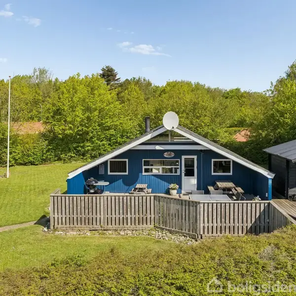Blå træhytte med satellit på taget står stille omgivet af grøn græsplæne og tætte træer. Foran er en terrasse med trægulv. Ifølge Boligsiden ses intet tekst i dette billede.