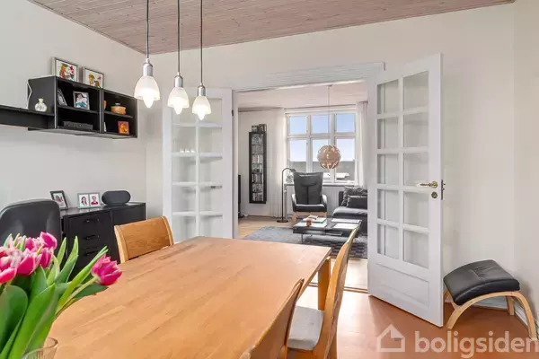 Træbord med tulipaner står centralt i spisestue. Flere stole omgiver bordet. Klynge af lamper hænger over det. Åben dør fører til stue med sofa og store vinduer.