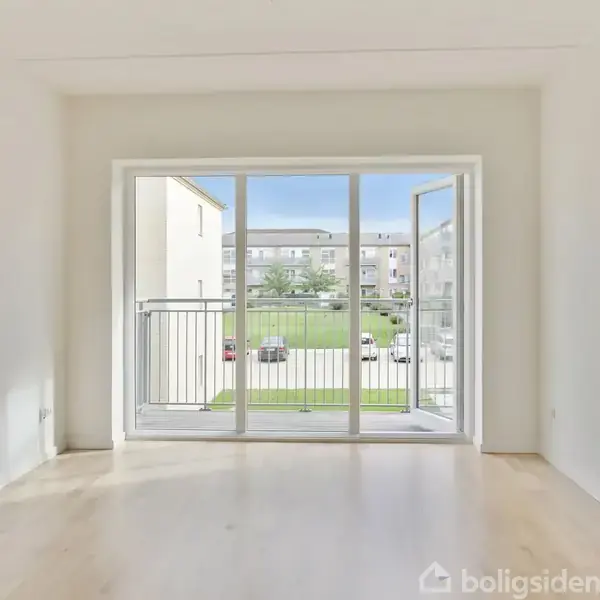 Et lyst rum med en stor åbentstående terrassedør, der fører ud til en balkon. Udsigt over en grøn gårdhave og lejlighedskompleks i baggrunden.