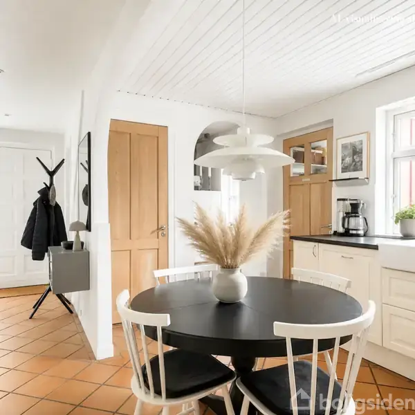 Spisebord med vase i køkken-alrum. Bordet har fire stole, en hvid lampe hænger over det. Køkkenet er lyst med hvide skabe, kaffemaskine og planter ved vasken. Entré ses til venstre.