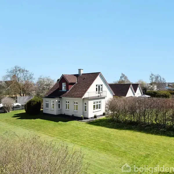En hvid, charmerende villa står stille på en velplejet grøn græsplæne omgivet af buske, under en klar blå himmel. En flagstang ses til venstre, og træer er i baggrunden.