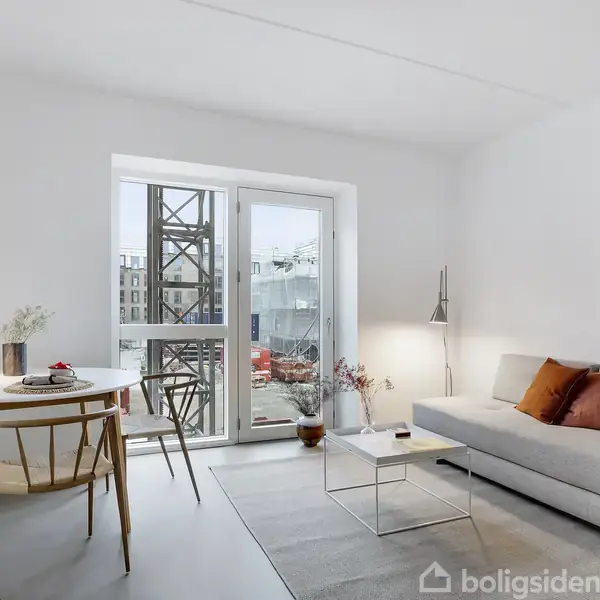 Stue med en lys grå sofa med farvede puder ved et glasbord. Der er et rundt spisebord med stole. Store vinduer åbner mod en byggeplads udenfor.