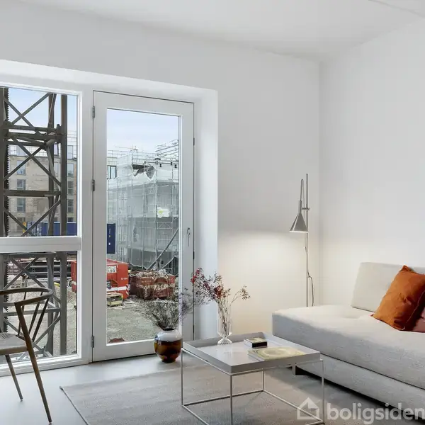 En moderne stue med en hvid sofa dekoreret med orange puder. En glasdør fører til en balkon med udsigt over en byggeplads. Et sofabord og en stålampe fuldender rummet.