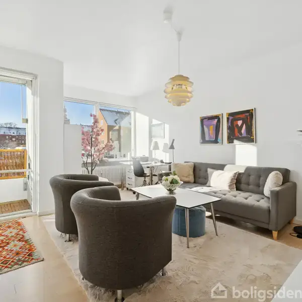 Stue med grå sofa og to stole, sofabord foran, og kunst på væggen. Lyst trægulv. Dør til balkon med et tæppe. Rummelig og lys med vinduer omkring. Ingen tekst forefindes.