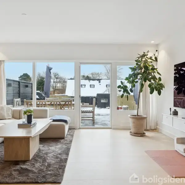 En lys stue med en stor sofa står ved et sofabord på et tæppe. Et vindue viser en snedækket terrasse. En plante står ved et TV på væggen.