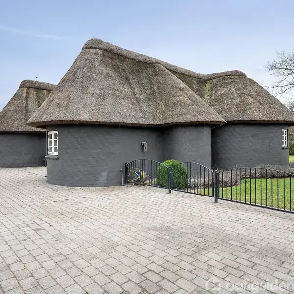 Et hus med stråtækt tag står stille på en flisebelagt gårdsplads, omgivet af grønne buske og træer mod en blå himmel.