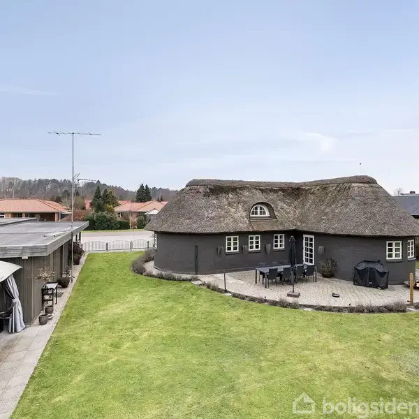 Stråtækt hus står i centrum af en velplejet have med græsplæne. Til venstre ses en pergola med hvide gardiner. Omkring ligger flere huse og der er en blå himmel.
