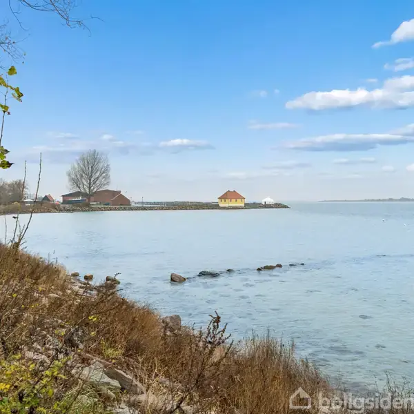 Kystlinje med små huse langs en rolig sø. Nogle træer og buske vokser tættere på kameraet, mens vandet strækker sig mod en fjern horisont under en blå himmel.