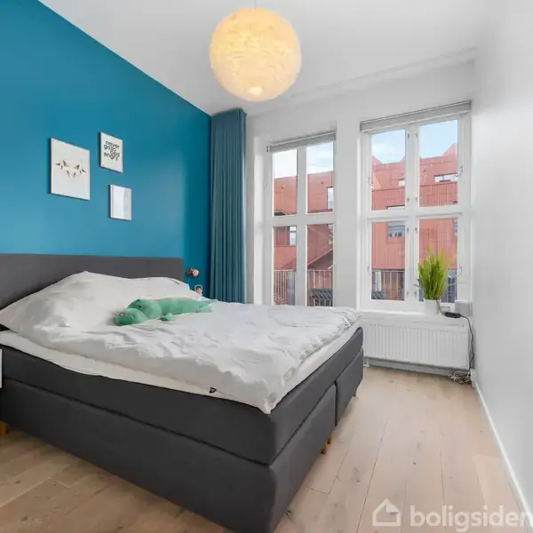 Soveværelset har en stor seng med gråt sengetøj, placeret mod en blå væg med kunstværker. Rummet er lyst med trægulv og et vindue, der viser røde bygninger udenfor.