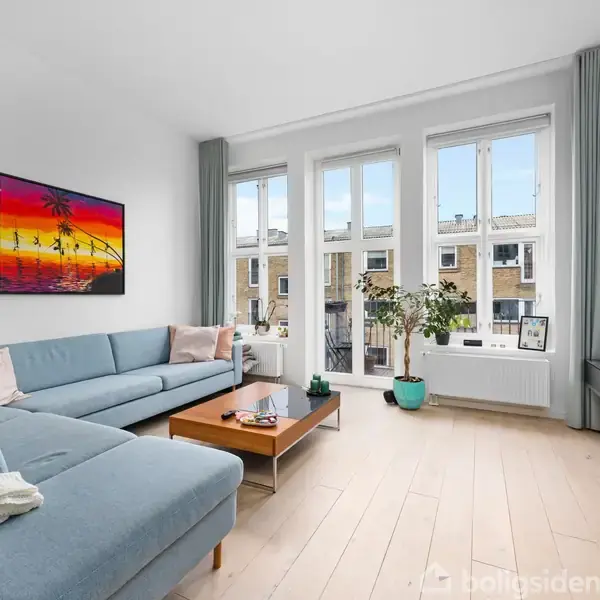 En lyseblå sofa er placeret i en stue foran et stort vindue med udsigt til bygninger. Der hænger et farverigt maleri på væggen, og en træbord står foran sofaen. Planter dekorerer rummet.