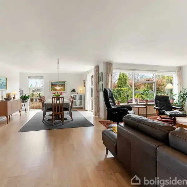 Stue med brune sofaer og et spisebord med stole centralt placeret på et tæppe. Store vinduer giver udsigt til en have. Møbler og planter pryder rummet.