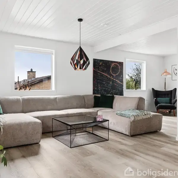Stor, beige hjørnesofa i en moderne dagligstue. Et sort stel-bord står foran sofaen, og en dekorativ lampe hænger fra loftet. Rummet er lyst med trælaminatgulv.