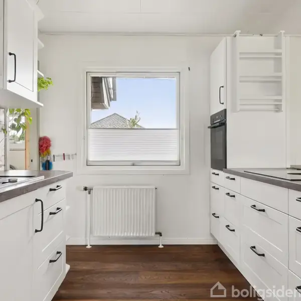 Hvidt køkken med sorte håndtag, trestatet vindue og runst radiator. Mørkt trægulv og vægmonteret ovn. Hængende lampe til højre, grønne planter på venstre hylde.