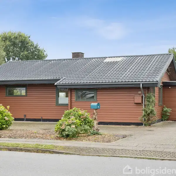 En brun træhus står stille på en villavej med en flisebelagt indkørsel. Der er buske foran huset og en garage på siden. Keine tekster er synlige.
