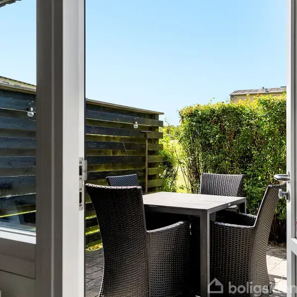 Dør åbner ud til en terrasse med et firkantet bord og fire fletstole. Baggrunden består af en hæk og et plankeværk.