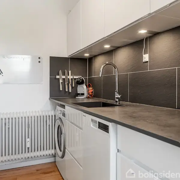 Køkken med hvidt interiør og grå bordplade. Knive hænger på væggen, opvaskemaskine og vaskemaskine under bordpladen. Væggen er beklædt med sorte fliser. Radiator og whiteboard til venstre.